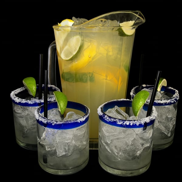 A pitcher of lemonade with lemon and lime slices, surrounded by four glasses of ice with lime wedges.