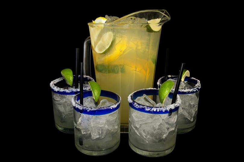 A pitcher of lemonade with lemon and lime slices, surrounded by four glasses of ice with lime wedges.