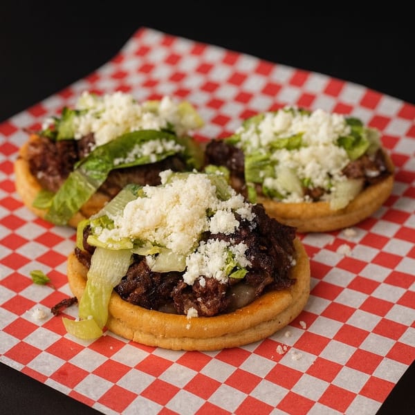 Three sopes with shredded meat, lettuce, and crumbled cheese on red and white checkered paper.