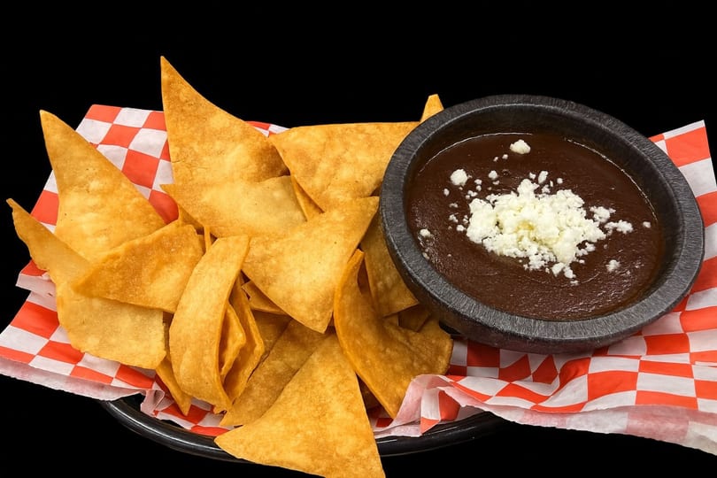 Basket of golden tortilla chips with a side of dark bean dip topped with crumbled white cheese.