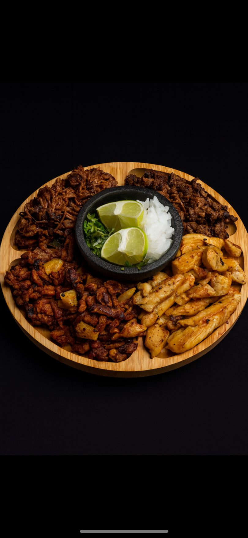 A wooden platter with four sections of different meats, a bowl of lime wedges, and diced onions.