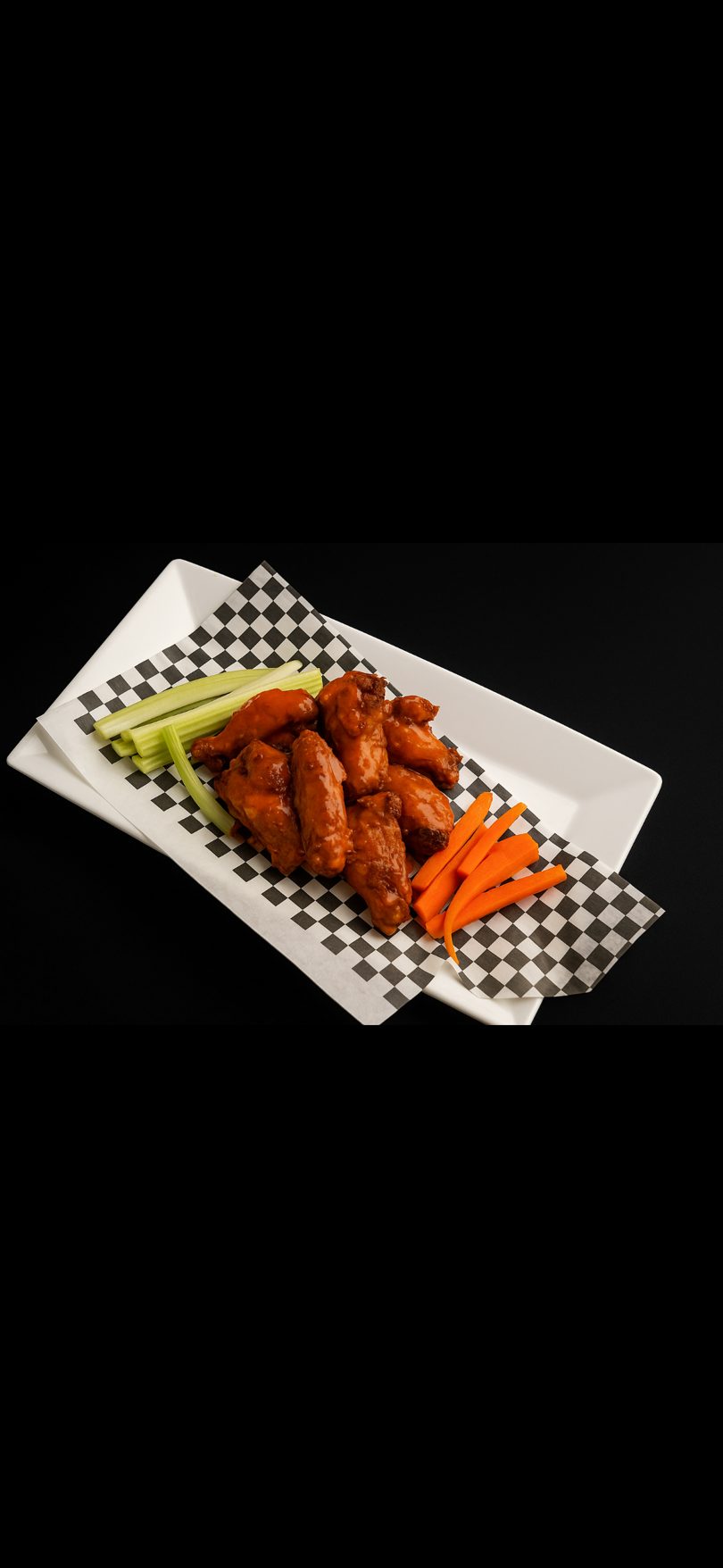 Spicy chicken wings with celery and carrot sticks on a white rectangular plate with checkered paper.