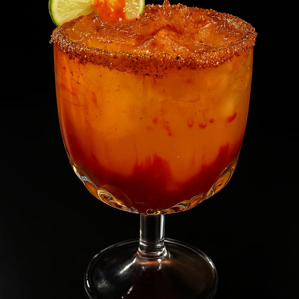 A vibrant orange drink with a chili-lime rim, garnished with a lime slice and red sauce.