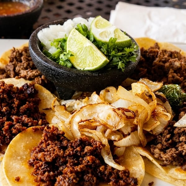 A plate of tacos with various meat fillings, grilled onions, a jalapeño, and a side of lime and cilantro.