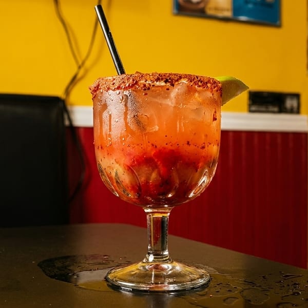 A vibrant red drink with a chili-lime rim and lime wedge, on a black table in a restaurant.
